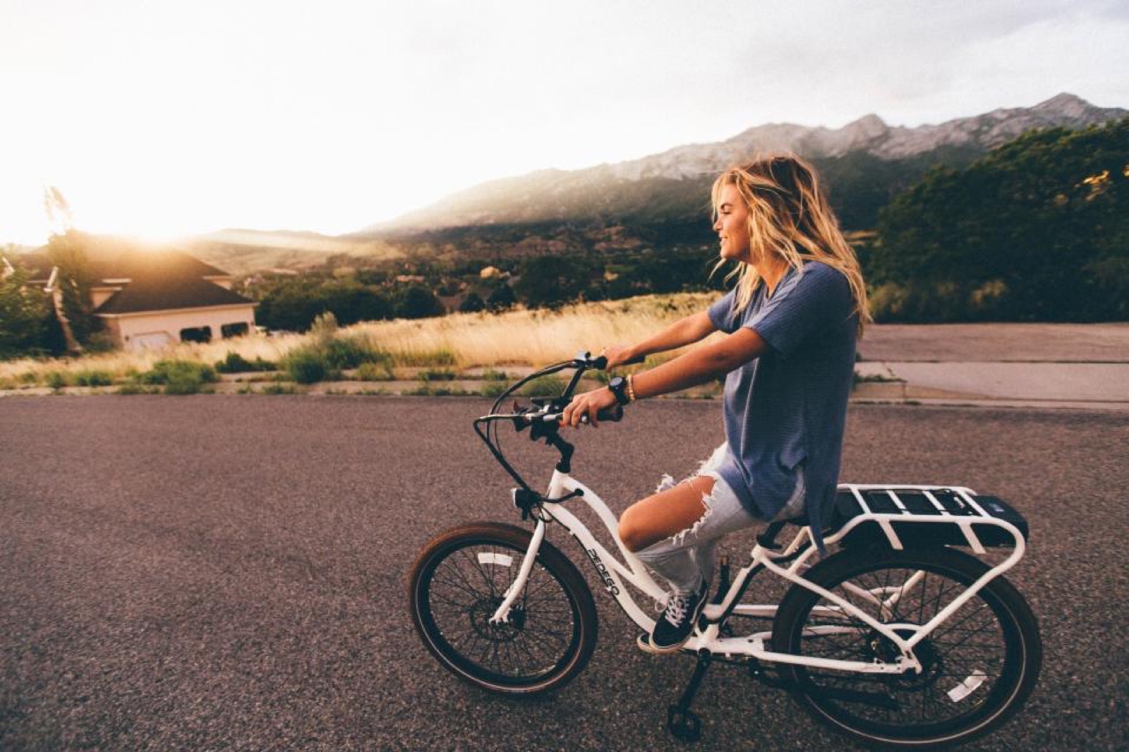 A rider on a white step-thru electric bike on a quiet road with mountains behind at sunset